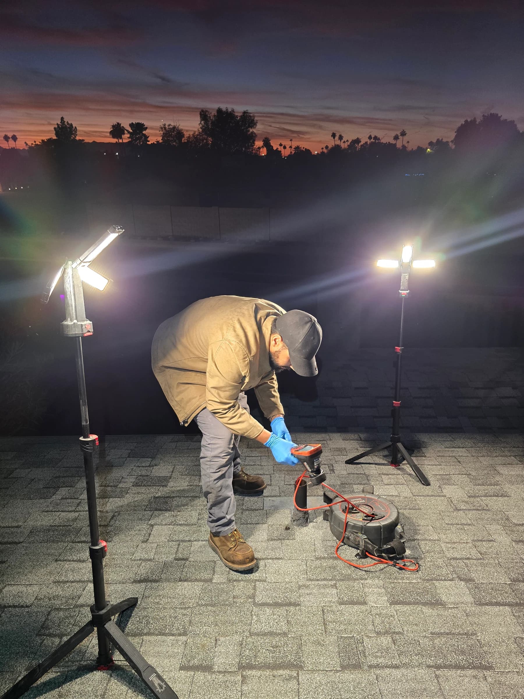Judai Brothers running a sewer camera from a roof vent at dusk, work lights on, palms behind the roofline